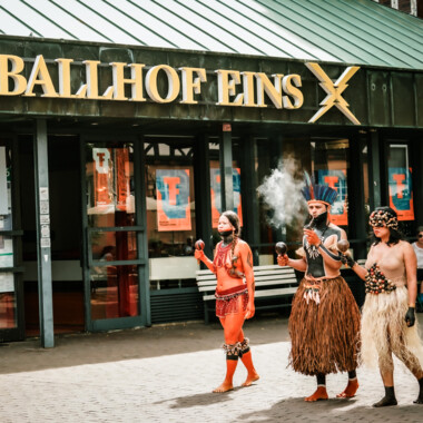 Drei Menschen in traditioneller Kleidung und Körperbemalung bei einer Prozession vor dem „Ballhof Eins“