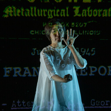 Chisato Minamimura in a white knee-length robe. To her left and right, she is bordered by two oversized green hands that are projected onto the dark wall behind her. She looks strained and, with her hands held up, gives the impression of wanting to push the projected hands away from her.