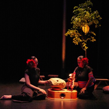 Zwei Personen sitzen auf einer Bühne. Sie tragen ein dunkles Kostüm und jeweils eine große rote Blume im Haar. Zwischen ihnen steht ein hölzernes Musikinstrument, dass an ein Grammophon erinnert. Anstelle einer Schallplatte liegt ein großer grauer Stein. Die beiden Personen scheinen das Instrument zu spielen.