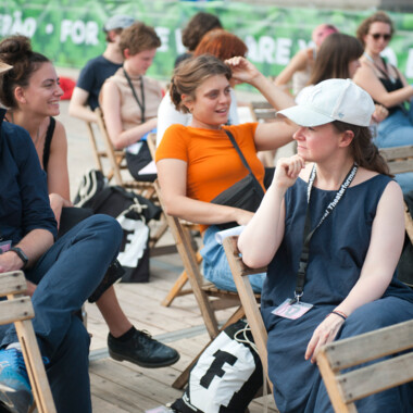  Festivalcampus-Teilnehmer*innen beim Festival Theaterformen. Sie sitzen in Holzstühlen. Rechts ein*e Teilnehmer*in mit weißer Cap und Festival Theaterformen-Lenyard in Schwarz. 