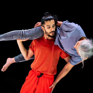 A dancing couple with a big age gap: Mohamed Toukabri and his mother Mimouna Latifa. Mohamed is wearing a tshirt and track pants in the color red. His black hair is tied up into a high ponytail. Across his shoulders is his mother, who is dressed in blue and has gray hair. He supports one of his mother's legs with his right hand, looking into her eyes. His mother's left shoulder rests on his left hand. Mimouna Latifa's left arm is outstretched, her hand touches Mohamed's thigh.