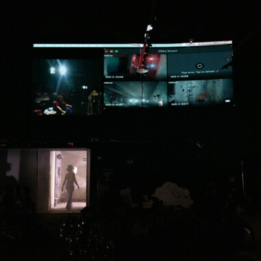 Ein dunkler Raum, oben werden auf einer großen Leinwand mehrere Live-Videobilder gezeigt werden. Darunter öffnet sich eine Tür, durch die eine Person als Silhouette in einen beleuchteten Raum tritt. Die Szene vermittelt eine kontrollierte, beobachtende Atmosphäre.