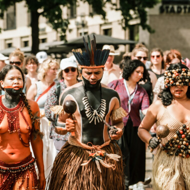 Drei Menschen in traditioneller Tracht führen eine Prozession durch eine Menschenmenge an.