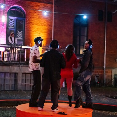 Vier Personen tanzen bei einer nächtlichen Silent Disco auf einem orangefarbenen Podest unter freiem Himmel. Sie tragen leuchtende Kopfhörer. Im Hintergrund steht eine Person an einem DJ Pult auf einem Balkon vor einer bunt beleuchteten Backsteinfassade.