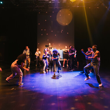 SPIN in der Cumberlandschen Bühne: Eine in blaues und gelbes Licht eingetauchte Tanzfläche, auf dem mehrere Personen im Kreis tanzen. Anna Seymour tanzt in der Mitte. 