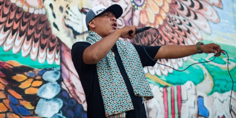 Wihntner FaGo during a hip hop show. Wihtner is wearing a black and white cap, a black T-shirt and a green patterned scarf over his shoulder. He caps the microphone in his right hand. Behind Wihtner is a colorfully painted wall.
