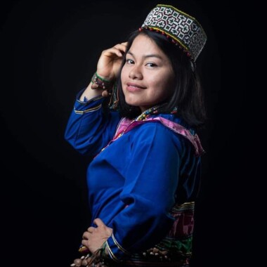 Chana Dávila in a blue dress with colorful appliqués. Chana has chin-length, dark, straight hair. She wears a patterned headdress on her head. Her right hand rests on her forehead, her left on her hip. She looks into the camera with a smile.