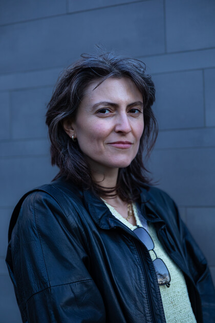 A portrait photo of Rita Mazza. Rita has black, shoulder-length hair with grey highlights. Rita is wearing a white T-shirt, a black leather jacket over it and has sunglasses tucked in front of the T-shirt. Rita is standing in front of a grey wall.