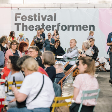 A lively scene at an outdoor festival with lots of people chatting, eating and drinking. A large banner with the words “Festival Theaterformen” can be seen in the background. The event takes place between a red brick building and a modern white building. There are colorful information stands, seating areas and a relaxed atmosphere.