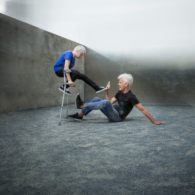 Self-description of the depicted persons: Two performers are in front of a gray stone wall and on gravel floor. On the right is Claire Cunningham, a short, white woman in her early forties.  She has short white hair and is wearing a blue t-shirt and black trousers and shoes.  Claire is leaning on crutches so that her feet are in the air. Her left foot is extended towards the person on the right. On the right is Jess Curtis, a tall man in his mid-fifties with an athletic build.  He has short white spikey hair and is wearing grey trousers and a black t shirt and shoes, which are dusty from the ground. Jess is sitting on the ground and touching the extended foot of the woman with his right hand. 