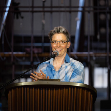 Anna Mülter during an opening speech. Anna has short gray hair, wears glasses and stands behind a microphone. She is wearing a blue and white shirt.