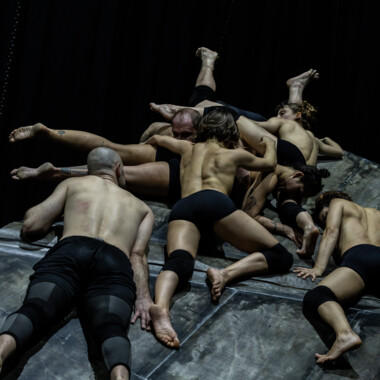 Mehrere Tänzer:innen liegen und klettern eng aneinander auf einer schrägen, rauen Metallfläche. Ihre Körper sind ineinander verschlungen. Alle tragen nur kurze schwarze Hosen und Knieschoner. Der Hintergrund ist dunkel.