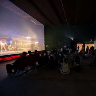 Publikum sitzt und steht in einem dunklen Raum und beobachtet eine Performance, die hinter einer transparenten Trennwand stattfindet. Die Szene auf der Bühne ist nur schemenhaft zu erkennen. Die Bühne ist warm beleuchtet, während der Zuschauerraum im Halbdunkel bleibt.