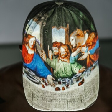 Top view of a person wearing a cap. A person with a bare back and arms outstretched to the side. The painting "The Last Supper" by Leonardo da Vinci is projected onto the cap.