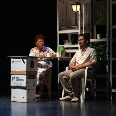 Eine ältere Frau und ein junger Mann sitzen auf einer Bühne an einem kleinen Tisch. Auf dem Tisch steht ein Wasserkocher und 2 Tassen. Neben ihnen stehen gestapelte Kartons, im Hintergrund Regale mit Alltagsgegenständen. Die Szene scheint wie eine häusliche Umgebung inszeniert.