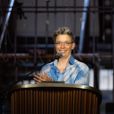 Anna Mülter während ihrer Eröffnungsrede beim Festival Theaterformen. Sie steht an einem Rednerpult und spricht in ein Mikrofon. Sie trägt ein blau-weißes Hemd, eine Brille und lächelt zum Publikum.