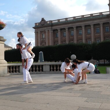 Mehrere Personen in weißen Kostümen performen auf einem öffentlichen Platz. Eine Person ist auf den Rücken einer anderen Person geklettert. Eine Person liegt am Boden, währen zwei weitere ihr hoch helfen zu wollen. Im Hintergrund ist ein historisches Gebäude.