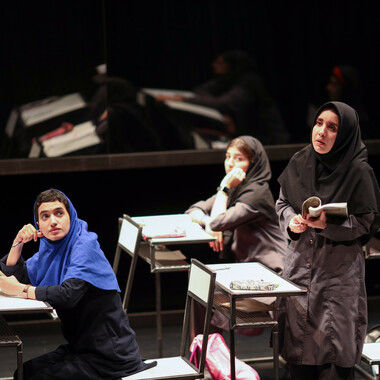 A classroom on the stage. On the right, a person is standing in a dark school uniform and black hijab. The person seems to be speaking while holding an open book in both hands. On the left, a female student in a blue hijab can be seen sitting at a desk and looking with wide eyes towards the other person speaking. In the background of the picture, another female student is sitting at the desk, her hair is covered with a black hijab. The person rests her head on her left hand.