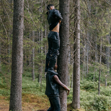 Eine Szene aus dem Stück “Undersang” von Harald Beharie: Es zeigt zwei Personen in schwarzen Lederklamotten in einem Wald. Eine Person davon umarmt einen Baumstamm, auf den Schultern steht die zweite Person. Die zweite Person hebt ihr Oberteil leicht hoch und berührt mit dem Bauch den Baumstamm.