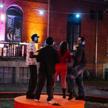 Vier Personen tanzen bei einer nächtlichen Silent Disco auf einem orangefarbenen Podest unter freiem Himmel. Sie tragen leuchtende Kopfhörer. Im Hintergrund steht eine Person an einem DJ Pult auf einem Balkon vor einer bunt beleuchteten Backsteinfassade.