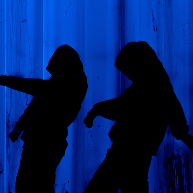 Bildbeschreibung seitens der Festival Theaterformen Redaktion. Selbstbeschreibung der abgebildeten Personen folgt. / Schwarze Silhoutten zweier Personen, die Kapuzenpullis tragen und tanzen, vor einem dunkelblauen Hintergrund.   