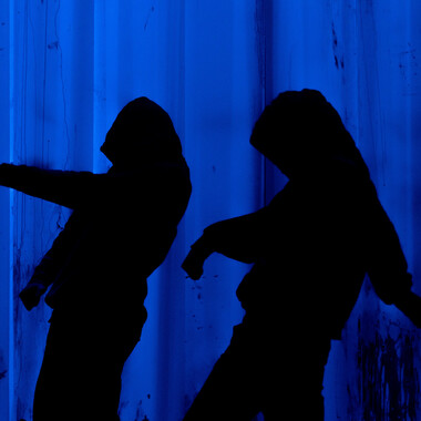 Bildbeschreibung seitens der Festival Theaterformen Redaktion. Selbstbeschreibung der abgebildeten Personen folgt. / Schwarze Silhouetten zweier Personen, die Kapuzenpullis tragen und tanzen, vor einem dunkelblauen Hintergrund.