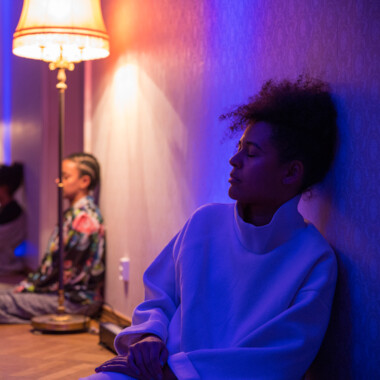 Mehrere Menschen sitzen entlang einer Wand in einem Raum mit warmem, gedämpftem Licht. Im Vordergrund lehnt eine Person mit geschlossenen Augen an der Wand.