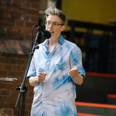 Anna Mülter during an opening speech. Anna has short gray hair, wears glasses and stands behind a microphone. She is wearing a blue and white shirt.