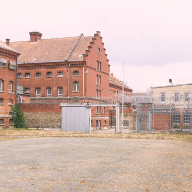 Ein Bild der JVA Rennelberg. Ein rotes Backsteingebäude mit vergitterten Fenstern, einen Hof mit Zäunen und Stacheldraht unter grauem Himmel.