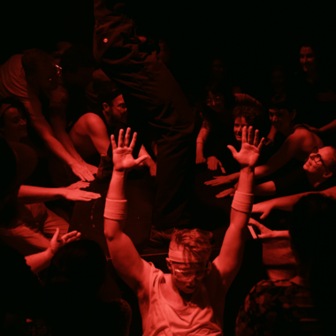 A club room immersed in red light, filled with guests. In the center of the lower half of the image, a person can be seen leaning backwards into other people and the subwoofer speaker, with his arms wide open. The person is wearing a white loose undershirt and large glasses. A wide white headband holds their short hairstyle. The person wears wrist sweatbands.