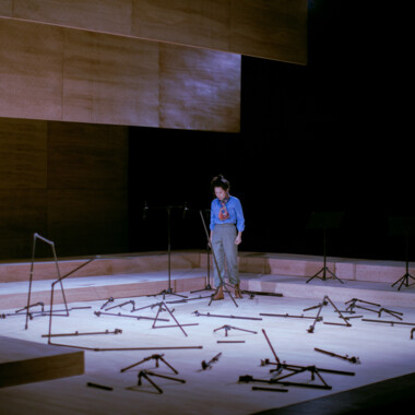 Eine Person steht allein auf einer Bühne, umgeben von verstreuten Metallgestellen und Stativteilen, die über den ganzen Boden verteilt sind. Die Person blickt auf zu Boden, als schaue sie sich die Szenerie an. 