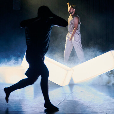 Bildbeschreibung seitens der Festival Theaterformen Redaktion. Selbstbeschreibung der abgebildeten Personen folgt. / Im Vordergrund ist die dunkle Silhouette einer Person zu sehen, die sich die Hände an den Kopf hält und rennt. Sie trägt ein T-Shirt und eine kurze Hose. Im Hintergrund stehen drei weiß-leuchtende Quader im Nebel und dahinter ist eine Person mit erhobener Faust und breitbeiniger Haltung, die nach links schaut. Die Person trägt eine weiße lange Hose und ein beiges Tanktop.