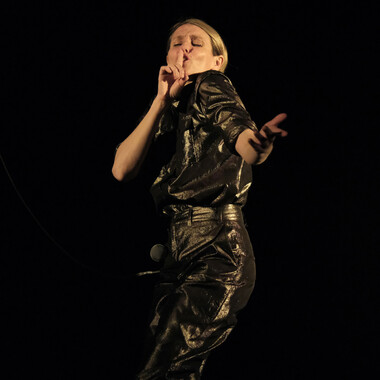 A darkened stage where Ivana Jozic performs in a short-sleeved dark leather jumpsuit. Ivana has pulled her short blonde hair into a deep side parting. With her eyes closed, Ivana holds her right index finger in front of her slightly open mouth. At the same time she stretches her left arm in a playful way forward and forms an indefinable gesture with her hand. The head of a microphone can be seen between Ivana's legs.