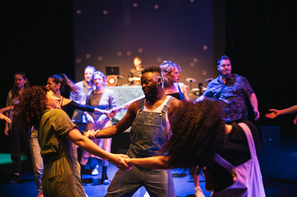 Szene aus SPIN in der Cumberlandschen Bühne: Eine in blaues Licht eingetauchte Tanzfläche. Anna Seymour (links), Chris Fonseca (Mitte) sowie Raffie Julien (rechts) im Bildvordergrund. Sie tanzen im Kreis und halten sich dabei an den Händen. Hinter ihnen sind weitere tanzende Menschen zu sehen.