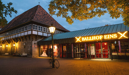 Das Ballhof Eins-Gebäude mit einem Schild mit der Aufschrift Ballhof Eins. Die gelbe Schrift ist hell erleuchtet. Das Schild befindet sich über den Eingang, der verglast ist. Vor dem Gebäude steht eine Laterne, die auch leuchtet.