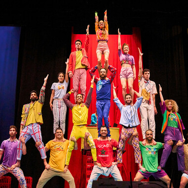 A five-tiered pyramid of colorfully dressed, happy people on stage, standing on each other's shoulders and thighs. Except for the bottom row and the person in the middle of the pyramid, everyone raises an arm in the air. In the background, colorful monochrome fabric panels can be seen.