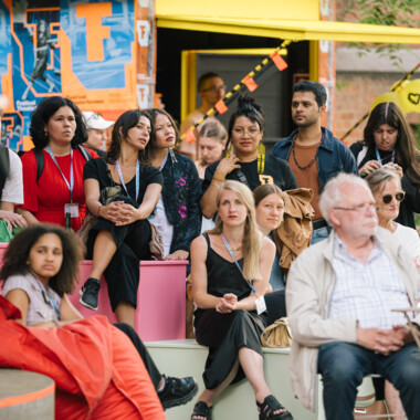 A group of people sit attentively on colored steps outside during an event. Orange and blue festival posters and a yellow canopy can be seen in the background. Some people are wearing festival passes around their necks. The atmosphere seems concentrated and interested.