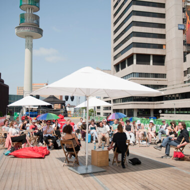 Students from the Festivalcampus Niedersachsen during a discussion session at the Theaterformen festival. They are sitting outside on wooden chairs, some of them under parasols. In the background is a high-rise building and a blue sky.