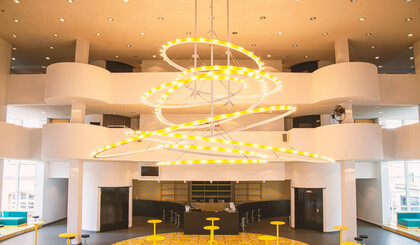  In the centre, a huge chandelier can be seen hanging from the ceiling of the foyer. It is characterised by abstract coils. The chandelier is striped yellow and white.