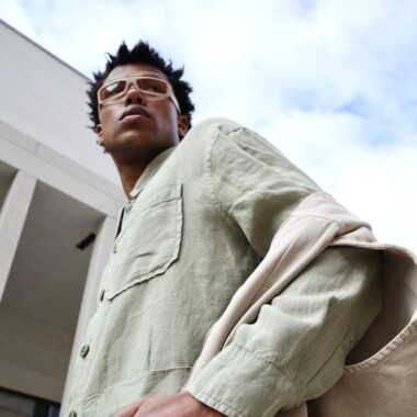 A young man in a mint green linen shirt looks confidently into the distance. He has a beige bag over his shoulder and is wearing clear-framed glasses. The photo is taken from a low angle, with a modern building and a lightly clouded sky in the background.