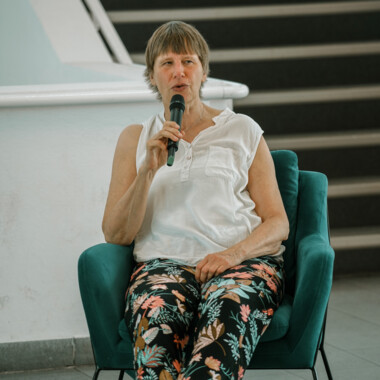 A person is seated in a turquoise armchair, holding a microphone in their right hand and speaking. They are wearing a sleeveless white top and black trousers with a colorful floral pattern. A staircase is visible in the background.