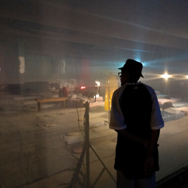 Eine Person mit Hut steht vor einer halbtransparenten Wand und blickt in einen Raum mit Matratzen, Lichtquellen und technischen Geräten. Die Szene ist diffus beleuchtet und nur schemenhaft zu erkennen.