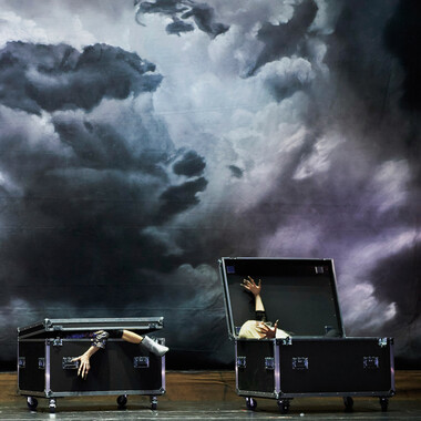 Two Flyht Cases with wheels, standing side by side on the stage like coffins. The left box is almost folded shut, an arm and a leg sticking out. The right box is unfolded and a vampire lying in the box stretches his arms out to either side. In the background there is a sky with dark clouds.