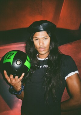 A man with long hair and a black beret holds a black bowling ball with green lettering "12" in his hand. He’s wearing a fitted black T-shirt with a white collar and a printed text across the chest. The red lighting in the background adds a dramatic effect to the scene.