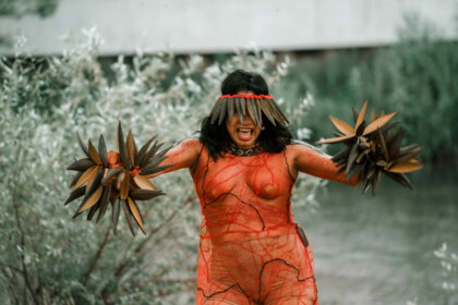 Eine Szenen aus der Performance Confluences, die an der Ihme stattgefunden hat. Die Performerin tragt Kopfschmuck der ihre Augen verdeckt, ihr Gesicht ist ausdrucksstark, ihr Mund geöffnet, als würde sie etwas rufen