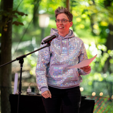 Anna Mülter während einer Eröffnungsrede. Anna hat kurze graue Haare, trägt eine Brille und steht hinter einem Mikrofon. In ihrer Hand hält sie ein Blatt Papier. Sie trägt einen grauen, glitzernden Kapuzenpullover und eine dunkle Hose. Hinter ihr Bäume.