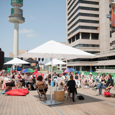 Studierende des Festivalcampus Niedersachsen während eines Gesprächsformat beim Festival Theaterformen. Sie sitzen draußen auf Holzstühlen, teilweiße unter Sonnenschirmen. Im Hintergrund ist ein Hochhaus und blauer Himmel.