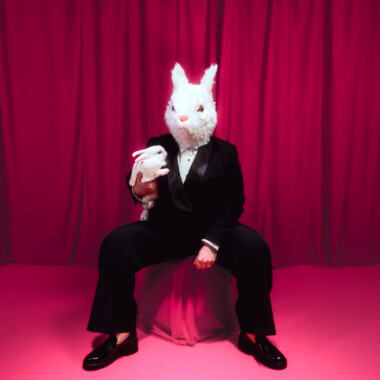 Eine Person in einem schwarzen Anzug sitzt auf Hocker sitzt. Sie trägt eine große, weiße Hasenmaske und hält ein weißes Stofftier in Form eines Hasen im Arm. Der Hintergrund besteht aus einem kräftig pinkfarbenen Vorhang und einem pinkfarbenen Teppich.