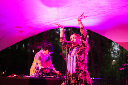 Hilda Länsman und Tuomas Norvio performen auf der Bühne im Festivalzentrum, die in pinkes Licht eingetaucht ist. Hildá Länsman singt und performt im Vordergrund, im Hintergrund Tuomas Norvio am Soundpult.