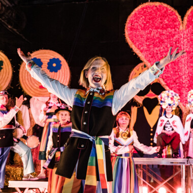 Stage setting: Lots of cheerful people in colourful traditional clothing, straw dolls and a huge heart in the background.